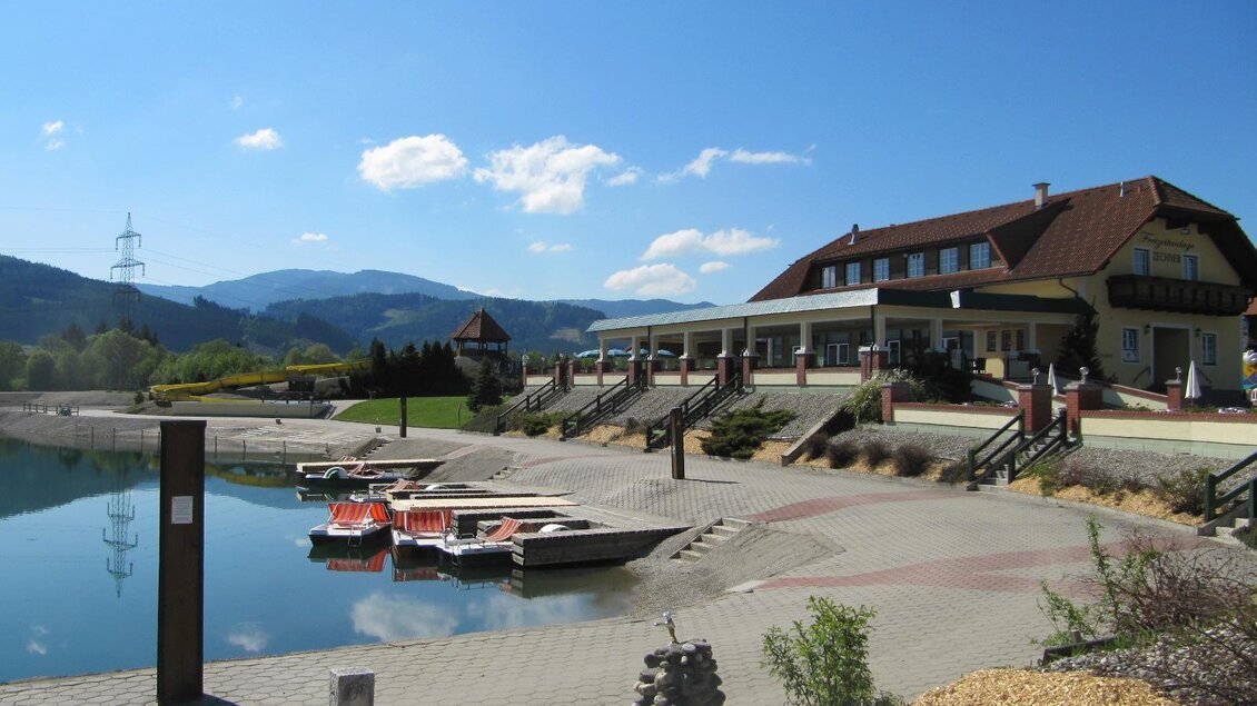 Ein ruhiger See mit Booten im Vordergrund und einem modernen Gebäude im Hintergrund. Im Hintergrund sind Hügel und ein blauer Himmel mit einigen Wolken zu sehen. | © Freizeitanlage Zechner