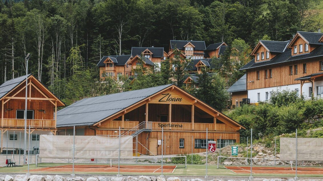 Eine malerische Sportarena mit Holzbau und Tennisplätzen umgeben von Bäumen. Im Hintergrund sind weitere alpine Gebäude zu sehen. | © BMI Austria