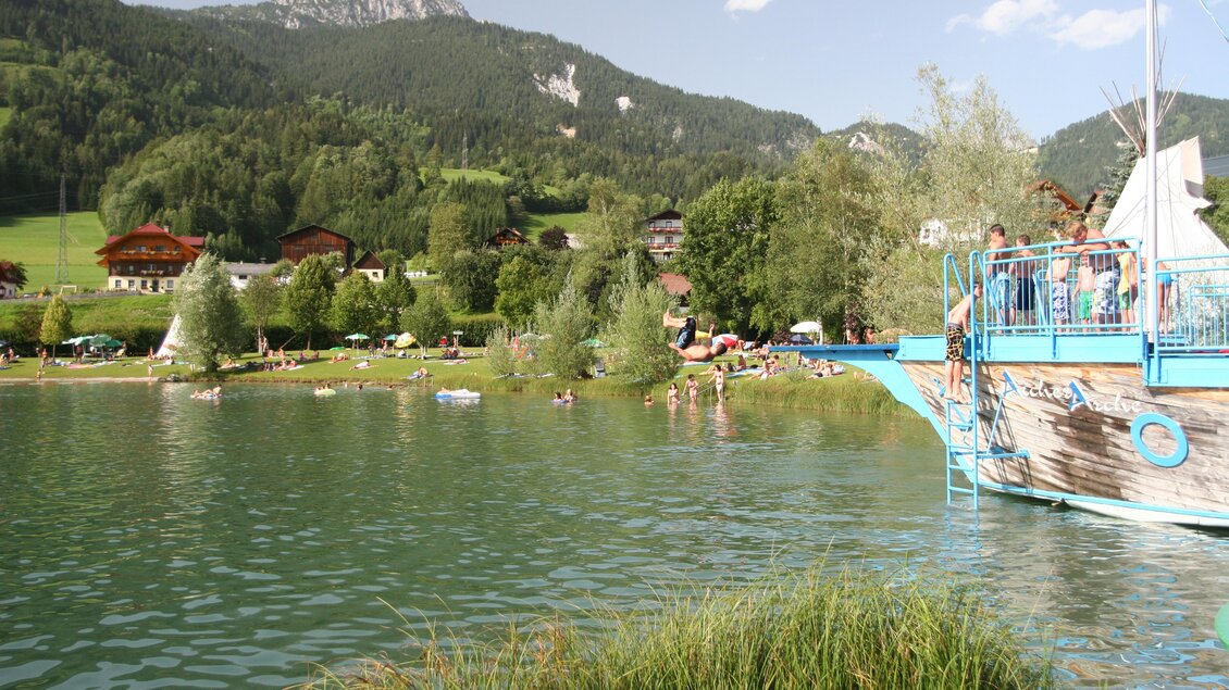 Ein ruhiger See mit klarem Wasser und einer grünen Umgebung. Im Hintergrund sind Berge und ein Bootsanleger zu sehen. | © Gemeinde Aich