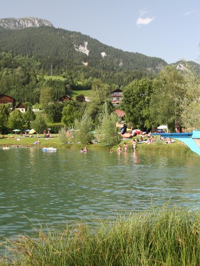 A peaceful lake with clear water and a green surrounding. In the background, there are mountains and a boat dock. | © Gemeinde Aich