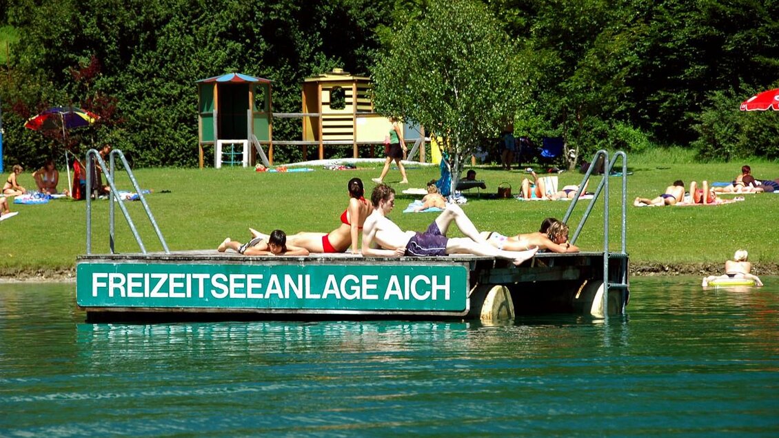 Ein Schwimmsteg am Wasser mit mehreren Leuten, die sich entspannen. Im Hintergrund ist eine grüne Wiese mit Spielgeräten und Sonnenschirmen. | © TVB Haus-Aich-Gössenberg