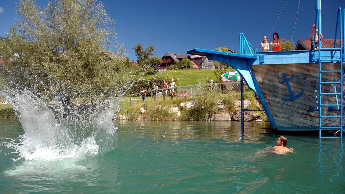 Ein Schwimmer im klaren Wasser springt von einem alten Holzboot und sorgt für Spritzer. Im Hintergrund sind grüne Wiesen und ein paar Menschen zu sehen. | © TVB Haus-Aich-Gössenberg