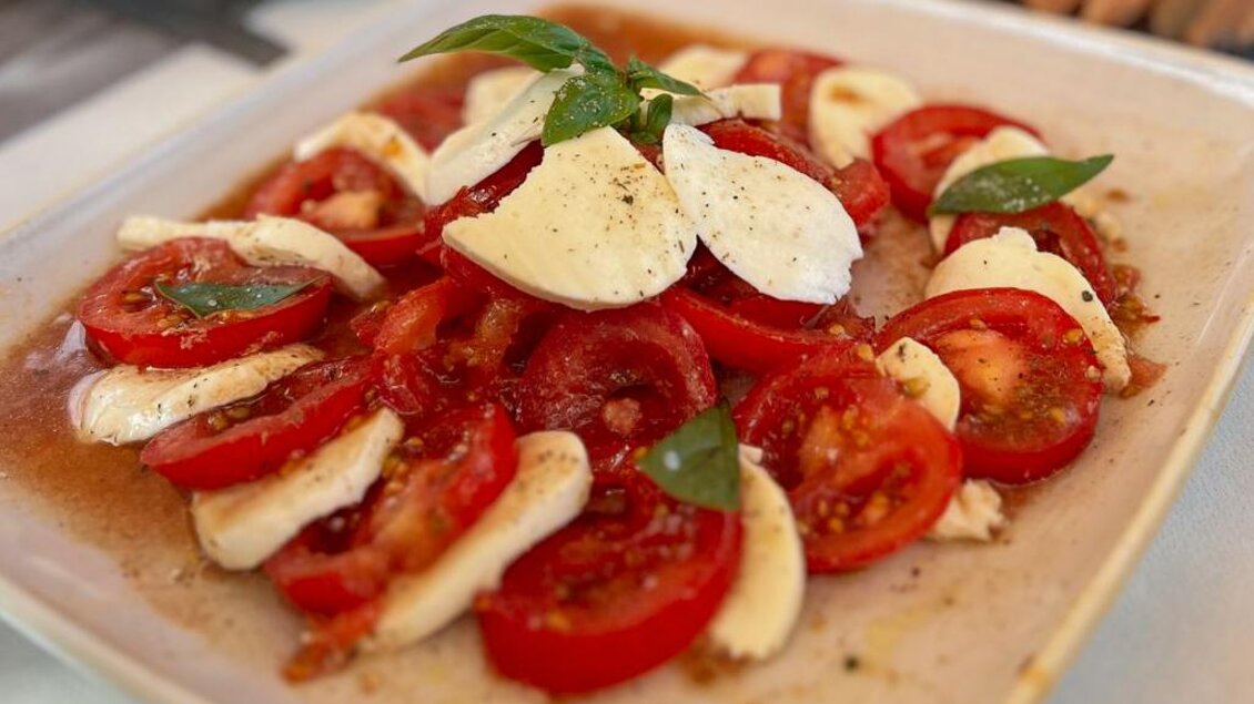 Ein Teller mit frischem Caprese-Salat, bestehend aus Tomatenscheiben, Mozzarella und frischem Basilikum. Das Gericht ist mit Olivenöl und Gewürzen verfeinert. | © Schuster Kulinarik OG