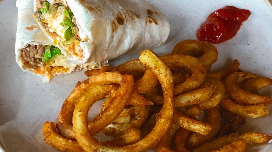 Ein Teller mit einem Wrap, gefüllt mit Fleisch, Gemüse und Käse, liegt neben einem Haufen gewellter Pommes frites und einer kleinen Menge Ketchup. Das Gericht sieht appetitlich aus und ist ideal für eine schnelle Mahlzeit. | © Schuster Kulinarik OG
