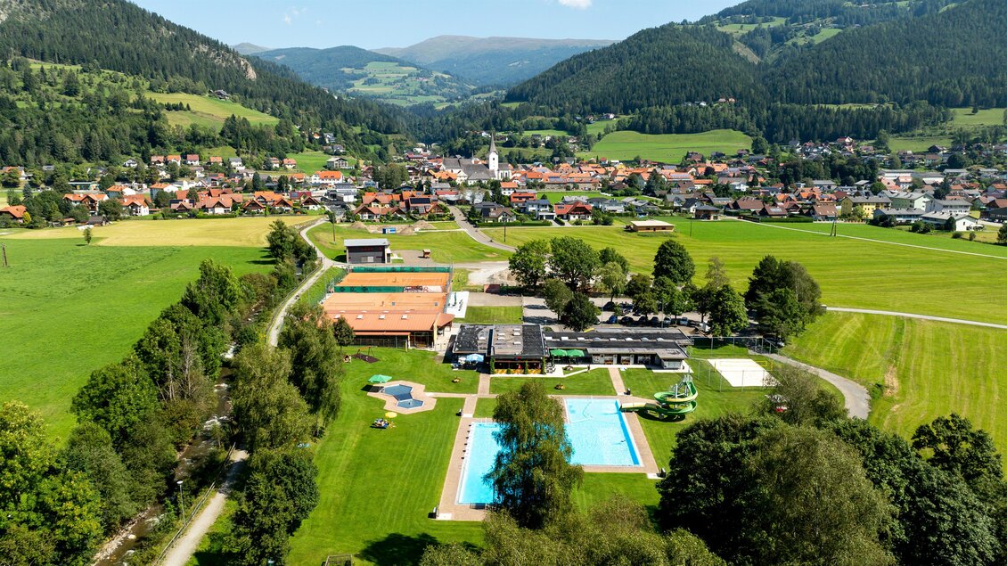 Eine ruhige Landschaft mit einem kleinen Dorf, umgeben von Hügeln und Wäldern. Im Vordergrund sind mehrere Pools und grüne Wiesen zu sehen. | © TV Murau