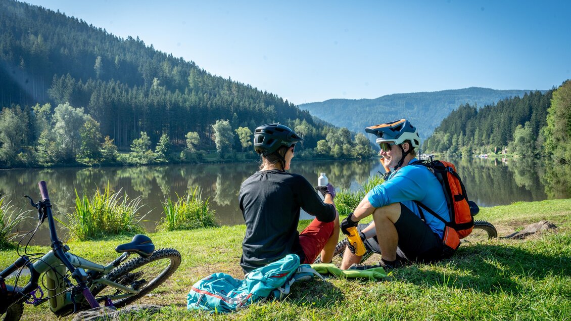 Zwei Radfahrer sitzen am Ufer eines ruhigen Sees und genießen die Landschaft. Im Hintergrund sind Wälder und Berge sichtbar. | © TV ERZBERG LEOBEN