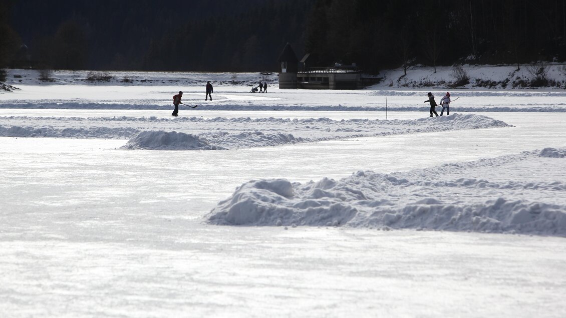 Eine winterliche Landschaft mit gefrorenem Wasser und Schneehügeln. Menschen genießen verschiedene Aktivitäten auf dem Eis. | © www.erzberg-leoben.at