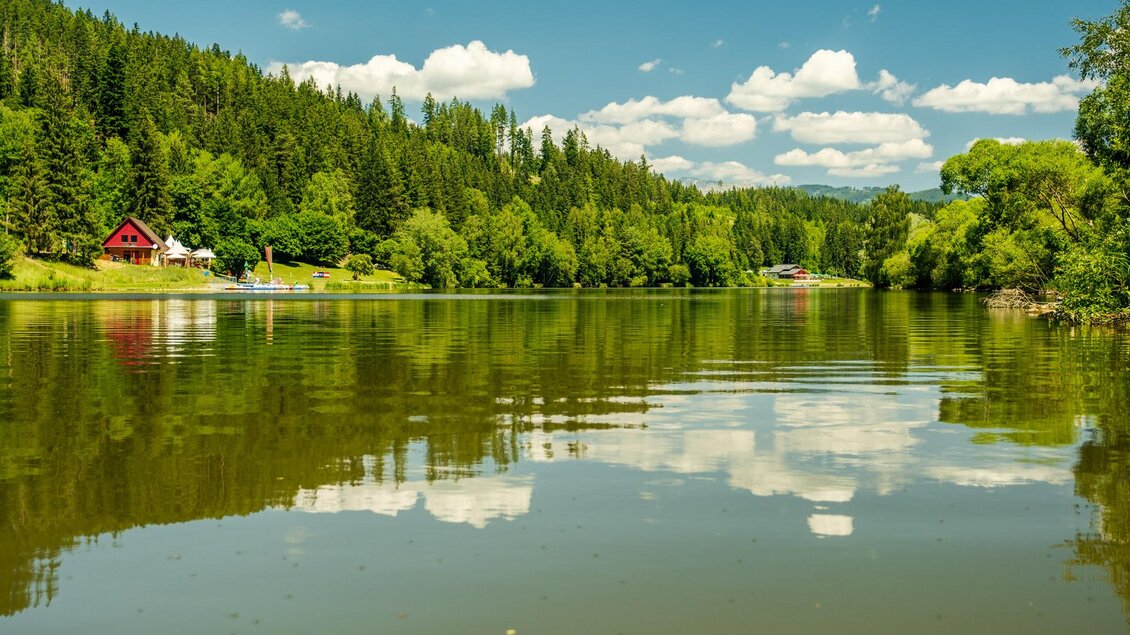 Ein ruhiger See umgeben von grünen Bäumen und Hügeln. Im Hintergrund ist ein rotes Holzhaus zu sehen, das sich im Wasser spiegelt. | © W. Spekner