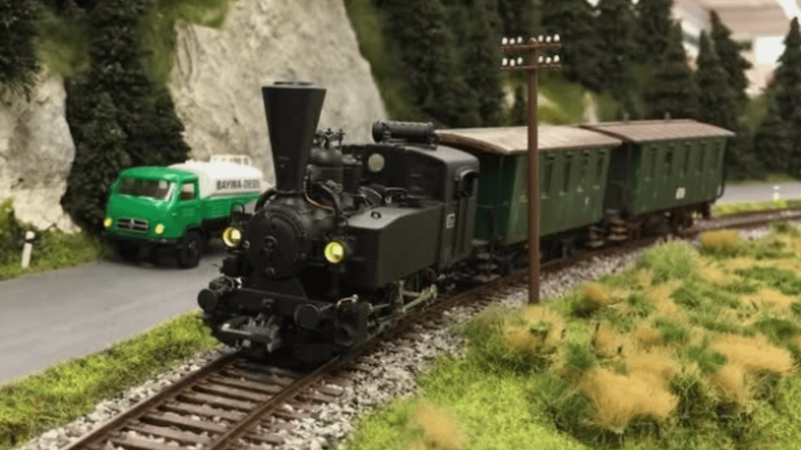 Eine alte Dampflok fährt auf einem Schienenweg entlang einer grünen Landschaft. Im Hintergrund ist ein Lkw und eine Stromleitung zu sehen. | © IG Freunde der Sulmtalbahn