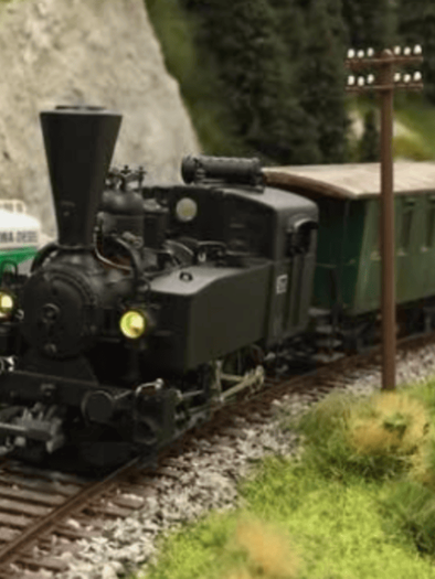 Eine alte Dampflok fährt auf einem Schienenweg entlang einer grünen Landschaft. Im Hintergrund ist ein Lkw und eine Stromleitung zu sehen. | © IG Freunde der Sulmtalbahn