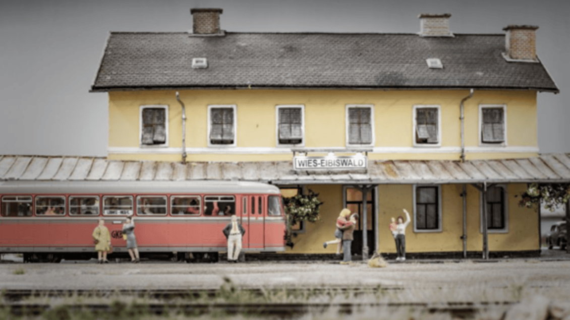 Eine Modellbahnhofszeile mit einem gelben Gebäude und einem roten Zug. Einige Figuren stehen auf dem Bahnsteig und scheinen aktiv zu sein. | © IG Freunde der Sulmtalbahn