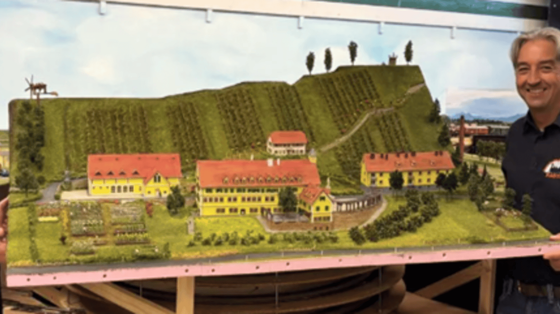 Zwei Männer halten ein detailliertes Modell eines Weinbaugebiets mit mehreren Gebäuden und einer hügeligen Landschaft. Im Hintergrund sind sanfte Hügel und ein blauer Himmel zu sehen. | © IG Freunde der Sulmtalbahn