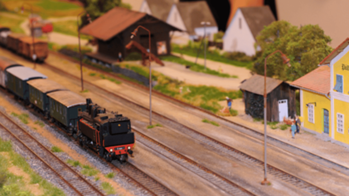 Eine Modellbahnanlage mit einer Dampflokomotive und mehreren Waggons. Im Hintergrund sind kleine Gebäude und Figuren zu sehen. | © IG Freunde der Sulmtalbahn