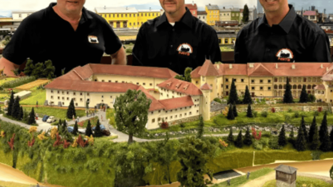 Drei Männer stehen vor einer beeindruckenden Modelleisenbahnanlage mit Landschaftsdetails und einem großen Gebäude im Hintergrund. Die Modelllandschaft zeigt Bäume, Wiesen und eine Gleisanlage. | © IG Freunde der Sulmtalbahn