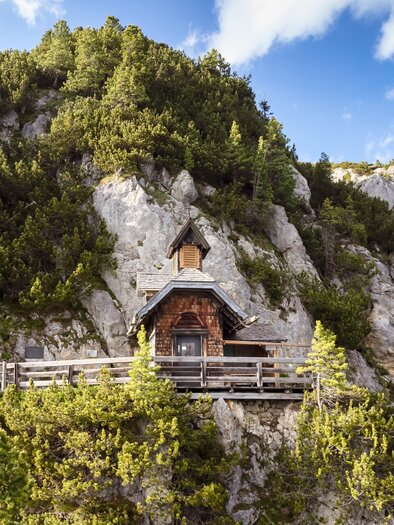 A charming small wooden house on a cliff, surrounded by green trees. In the background, impressive rocks and a blue sky can be seen. | © Schöttl/ORF