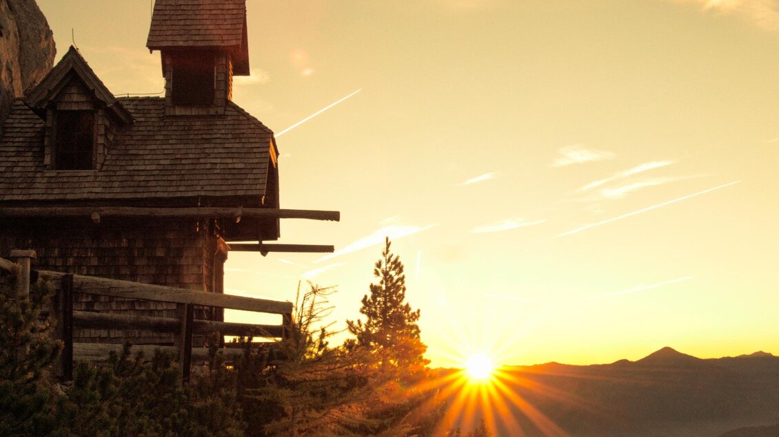 Ein malerisches Haus auf einem Hügel mit Blick auf die Berge bei Sonnenaufgang. Die Sonne strahlt durch die Äste der Bäume und taucht die Landschaft in warmes Licht. | © Erlebnisregion Schladming-Dachstein