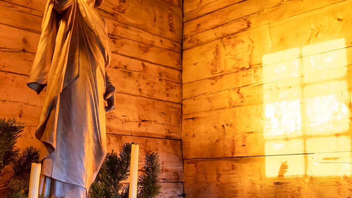 Eine Statue von Jesus steht in einem rustikalen Raum mit Holzwänden. Das Licht der untergehenden Sonne strömt durch ein Fenster und beleuchtet die Szene. | © Schöttl/ORF