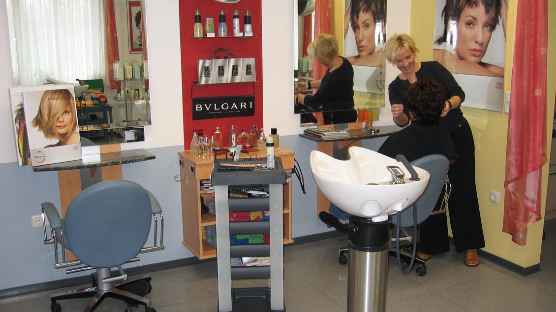 Ein Friseursalon mit zwei Stühlen und einem Waschbecken. An den Wänden sind Bilder von Haarschnitten und Beautyprodukten sichtbar. | © Friseur Ruck