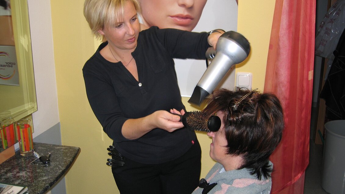 Eine Friseurin stylt das Haar einer Kundin mit einem Föhn. Im Hintergrund ist ein großes Bild einer anderen Frau zu sehen. | © Friseur Ruck