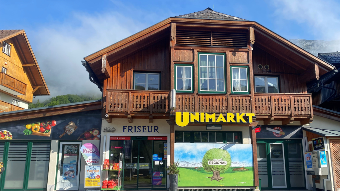 Ein traditionelles Holzgebäude mit Geschäften im Erdgeschoss. In der Mitte befindet sich ein UNIMARKT, links ein Friseur. | © Bettina Scheck