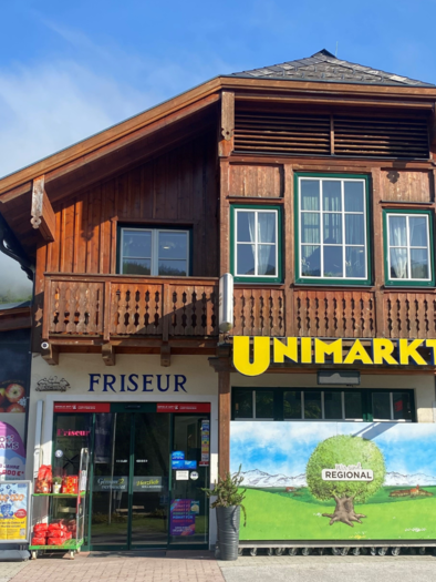 Ein traditionelles Holzgebäude mit Geschäften im Erdgeschoss. In der Mitte befindet sich ein UNIMARKT, links ein Friseur. | © Bettina Scheck