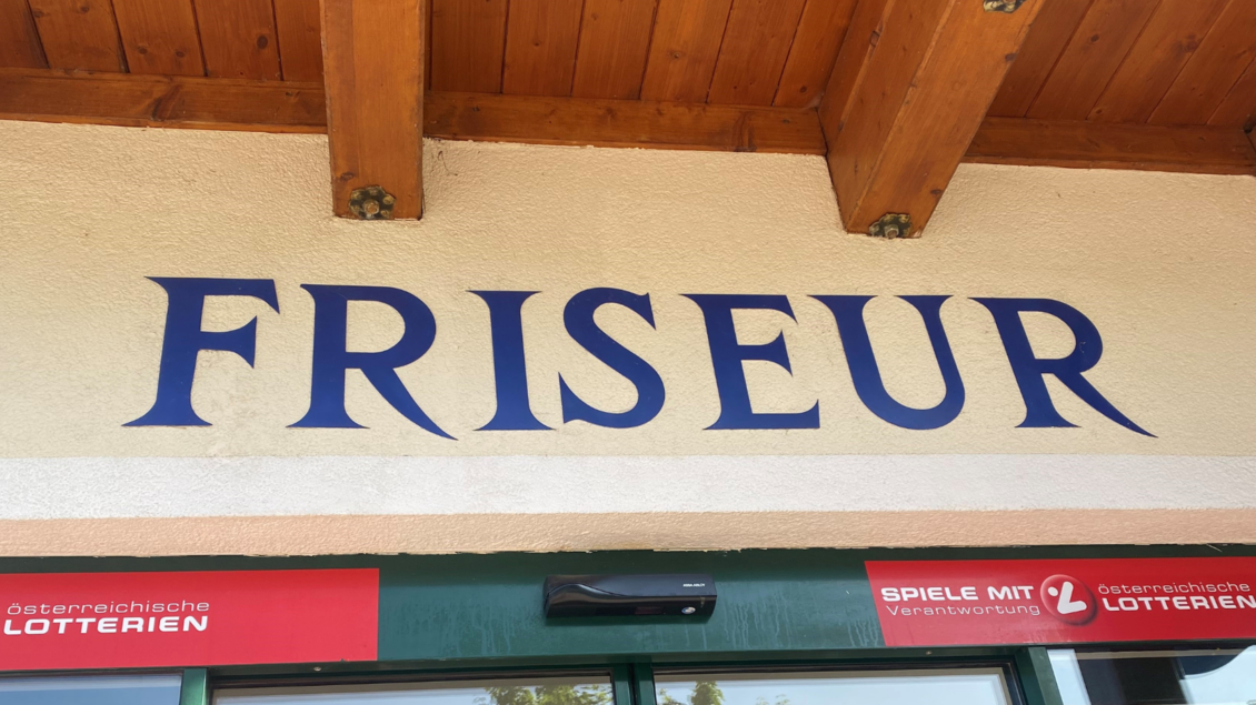 Ein Friseursalon mit dem großen Schriftzug „FRISEUR“ an der Wand. Darunter sind Werbeelemente für Lotterien sichtbar. | © Bettina Scheck