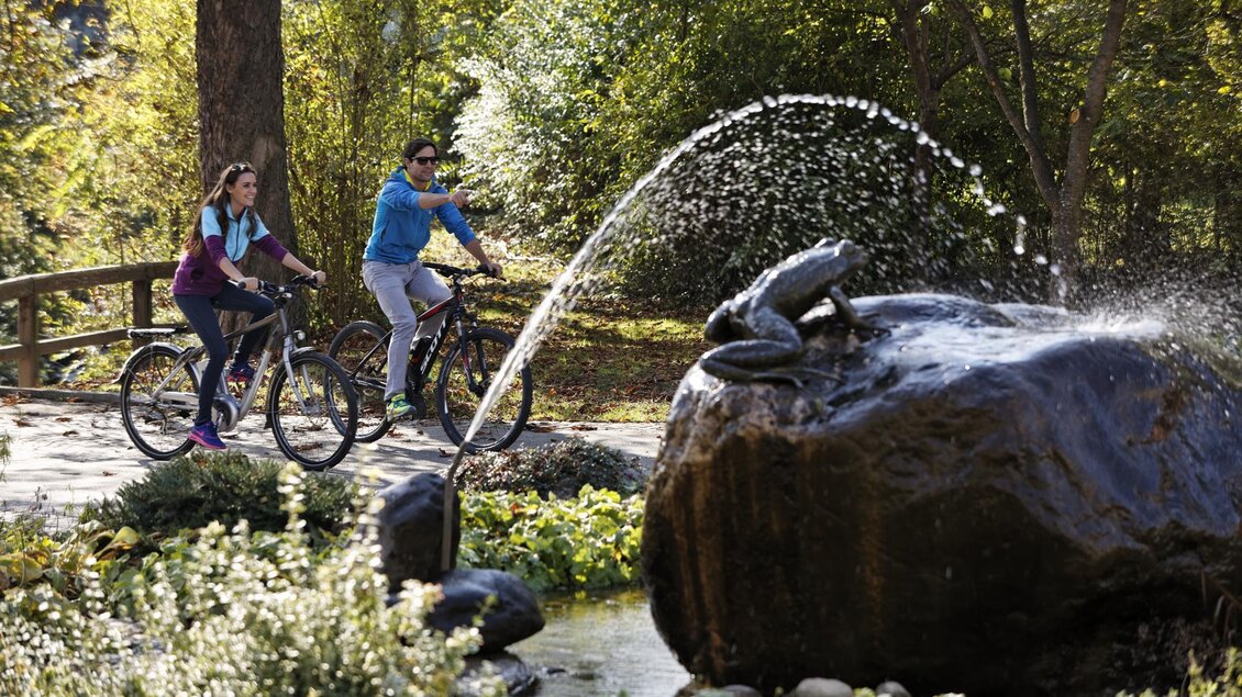 Zwei Radfahrer genießen einen sonnigen Tag in einem Park. Im Hintergrund springt Wasser aus einem Brunnen, der eine Froschfigur zeigt. | © Bernhard Bergmann
