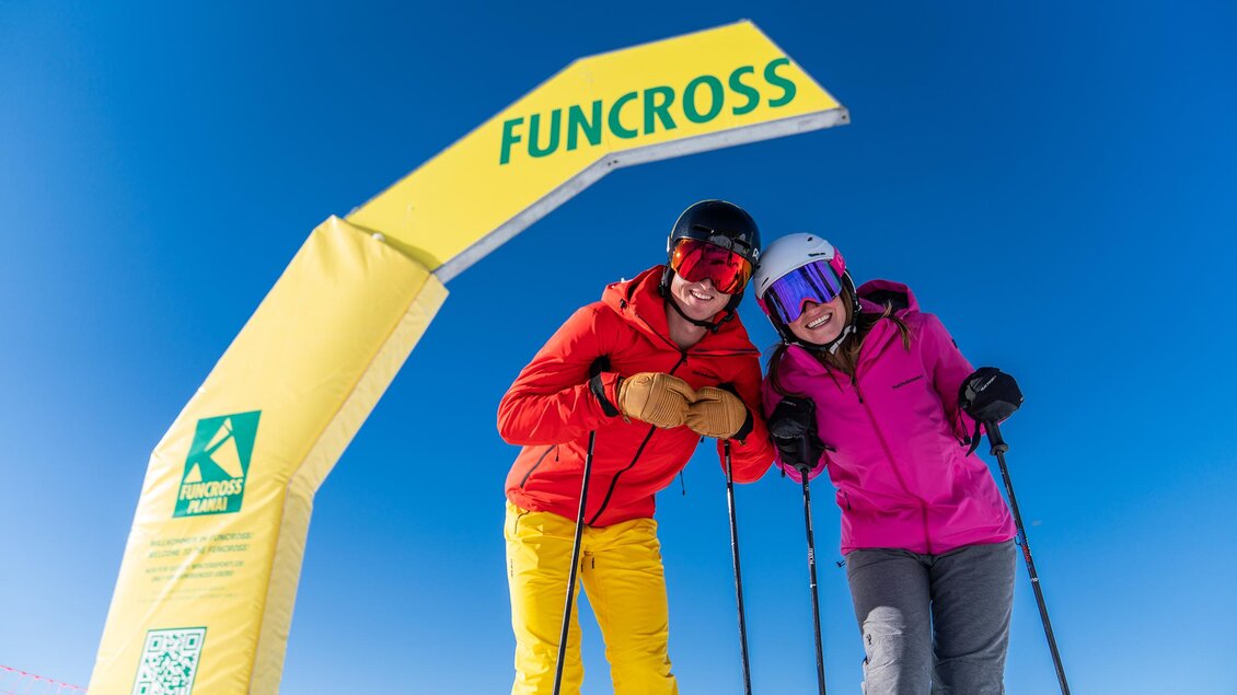 Zwei Skifahrer stehen unter einem gelben Funcross-Bogen in einer sonnigen Berglandschaft. Der Himmel ist klar und blau, was perfekte Bedingungen zum Skifahren verspricht. | © Christine Höflehner