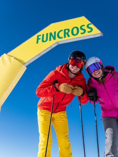 Two skiers are standing under a yellow Funcross arch in a sunny mountain landscape. The sky is clear and blue, promising perfect conditions for skiing. | © Christine Höflehner