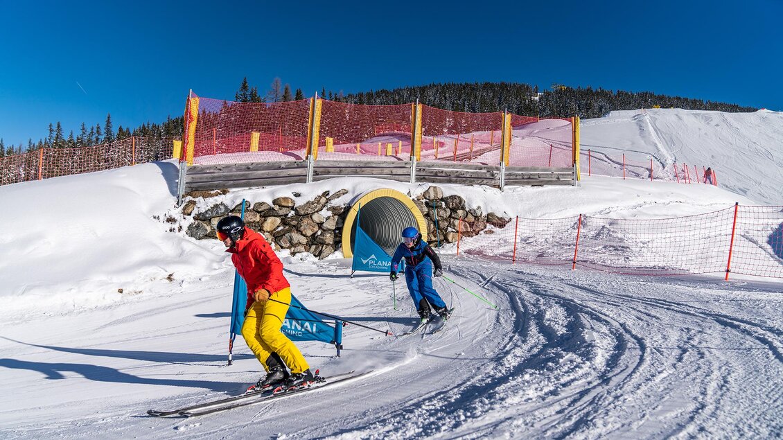 Zwei Skifahrer fahren eine Piste hinunter, umgeben von Schnee und einer klaren blauen Himmel. Im Hintergrund sind Sicherheitsnetze und ein Tunnel sichtbar. | © Christine Höflehner