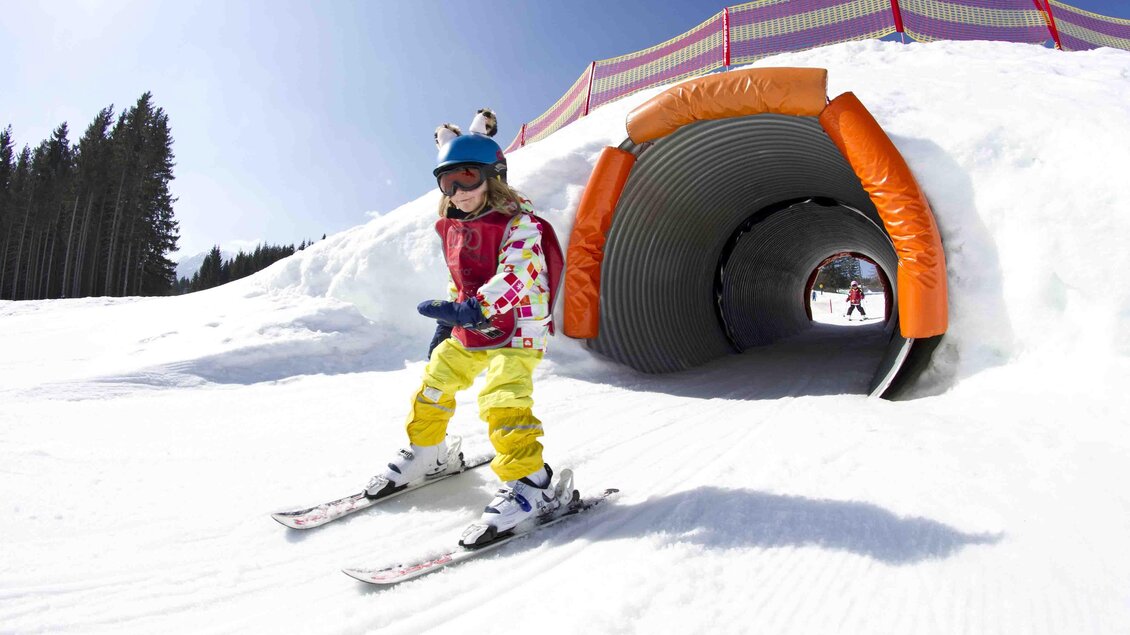Ein fröhliches Kind fährt auf Skiern und gleitet durch einen Tunnel aus Schnee. Im Hintergrund sind Bäume und blauer Himmel zu sehen. | © Katja Pokorn