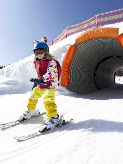 A happy child is skiing and gliding through a tunnel of snow. In the background, there are trees and a blue sky. | © Katja Pokorn
