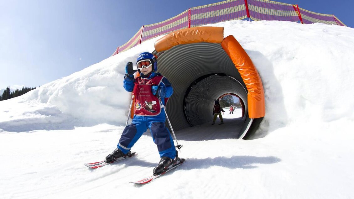 Ein fröhliches Kind in Skiausrüstung steht vor einem Tunnel im Schnee. Der Himmel ist klar und blau, ideal für einen Tag auf der Piste. | © Katja Pokorn