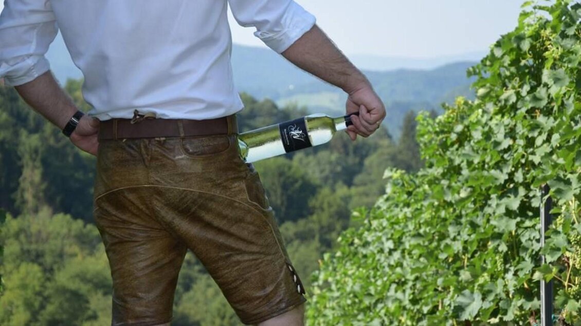 Ein Mann in Lederhosen hält eine Flasche Wein in der Hand. Im Hintergrund sind Weinreben und eine hügelige Landschaft zu sehen.
