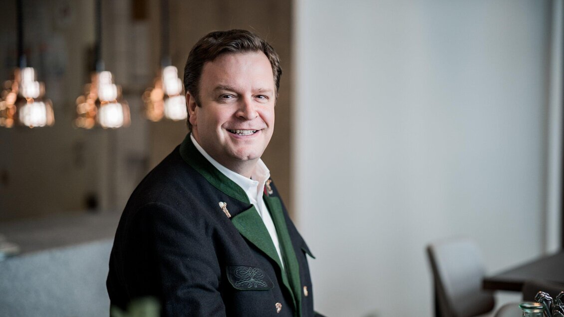 Ein lächelnder Mann in traditioneller Tracht sitzt in einem modernen Innenraum. Im Hintergrund sind stilvolle Lampen und Möbel zu sehen. | © Gösser Bräu - Tina Szabo