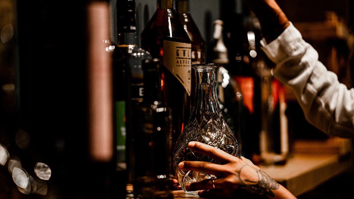 Eine Person greift nach einer eleganten Glaskaraffe vor einer Auswahl an Spirituosen. Die Beleuchtung schafft eine gemütliche Atmosphäre in einer Bar. | © Gösser Bräu - Tina Szabo