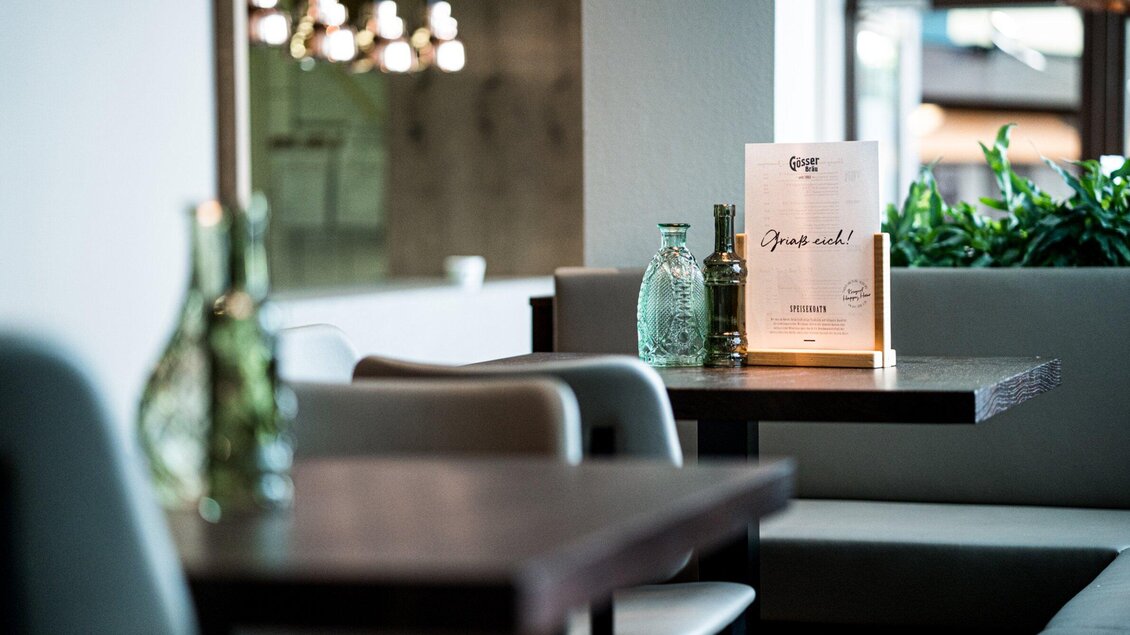 Ein gemütlicher Restaurantbereich mit Holz- und Tischdekor. Auf den Tischen stehen Wasserflaschen und eine Speisekarte. | © Gösser Bräu - Tina Szabo