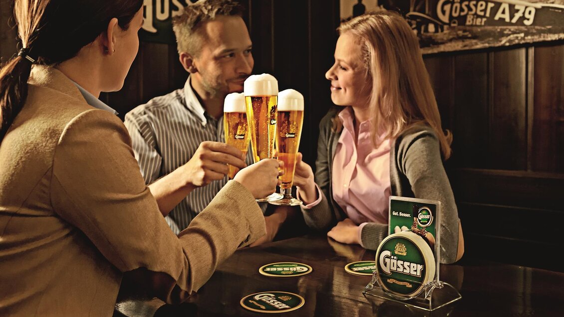 Drei Personen stoßen mit Biergläsern an. Im Hintergrund sind Getränkekarten und ein gemütliches Ambiente zu sehen. | © Brauunion