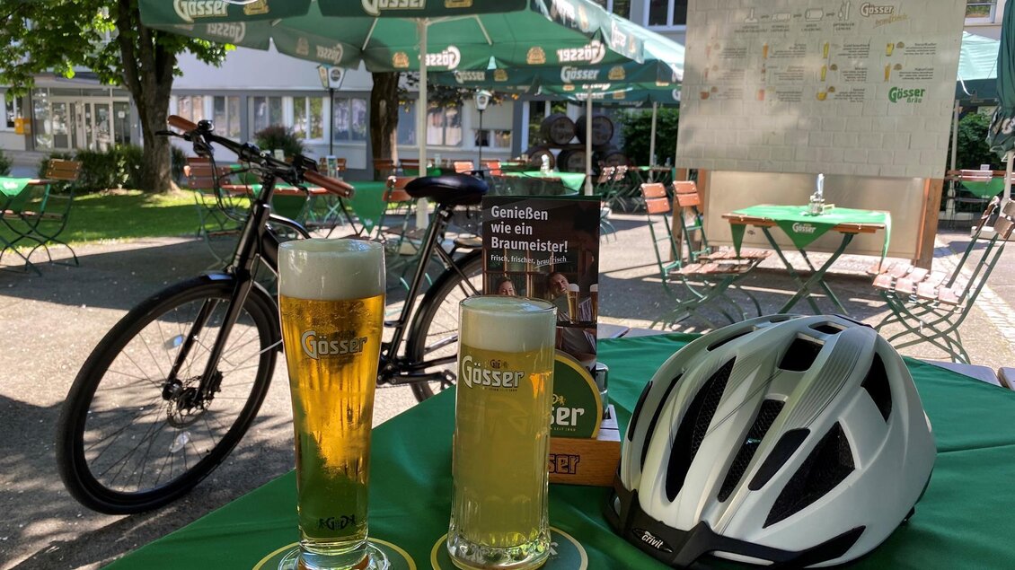 Zwei Gläser Bier stehen auf einem Tisch, umgeben von einem grünen Tischset. Im Hintergrund sind Fahrräder und Sonnenschirme zu sehen. | © Gösserbräu Leoben