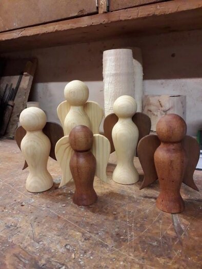 Six handcrafted wooden angels are standing on a table. They are made from different types of wood and colors.