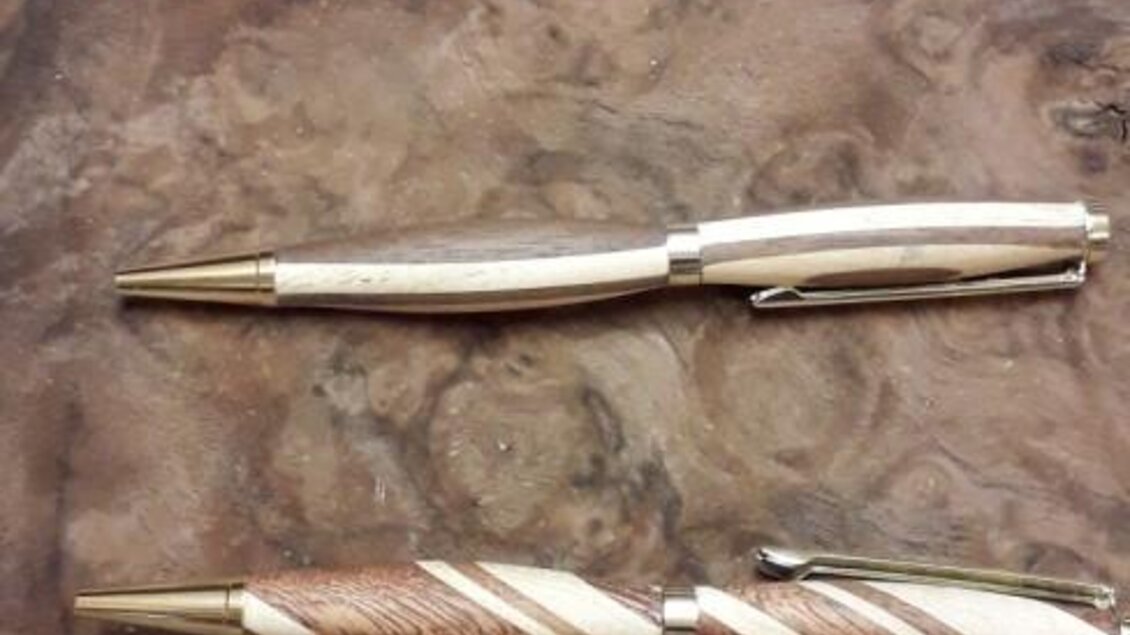 Zwei hochwertige Kugelschreiber liegen auf einer Holzoberfläche. Der obere Stift hat ein schlichtes Design, während der untere Stift eine gedrehte Musterung aufweist.