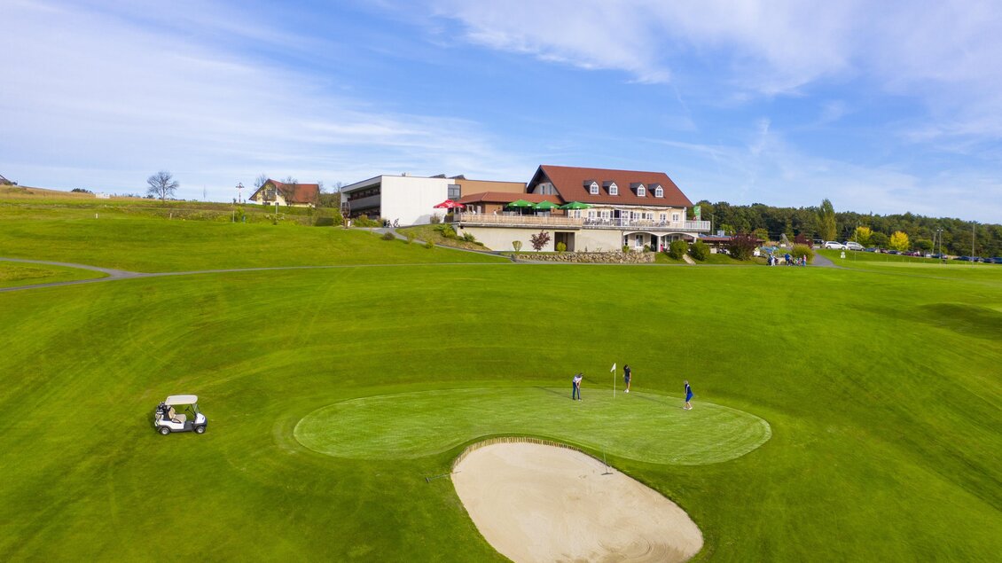 Eine weitläufige Golfanlage mit einem Clubhaus im Hintergrund. Auf dem Grün befinden sich Golfer, die die Anlage nutzen. | © TV- Thermen- & Vulkanland