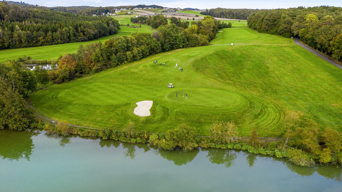 Eine grüne Golfanlage mit mehreren Spielbahnen und einem kleinen Wasserhindernis. Bäume umranden das Gelände und die Landschaft ist weitläufig. | © TV- Thermen- & Vulkanland