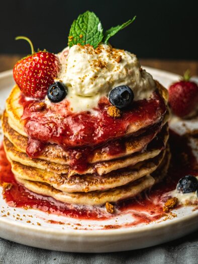A stack of fluffy pancakes topped with fresh strawberries and blueberries. The pancakes are adorned with a fruity sauce and a creamy topping. | © Granola