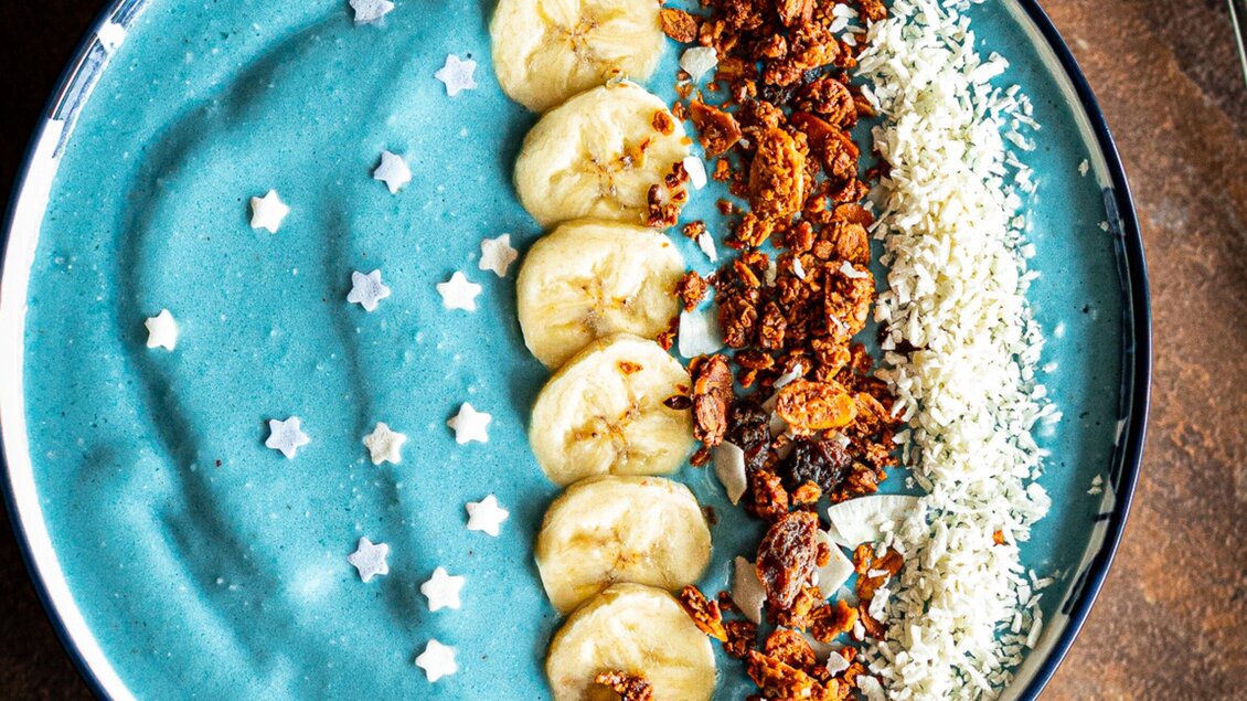 Eine farbenfrohe Schüssel mit blauer Smoothie-Basis, garniert mit Bananenscheiben, Granola und Kokosraspeln. Kleine weiße Sterne verleihen dem Gericht eine festliche Note. | © Granola