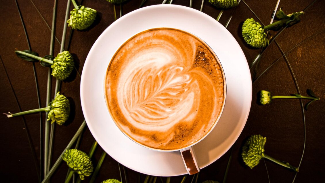 Eine Tasse Kaffee mit kunstvollem Milchschaum liegt auf einem Teller. Rundherum sind grüne Pflanzen und die Tasse ist schön dekoriert. | © Granola