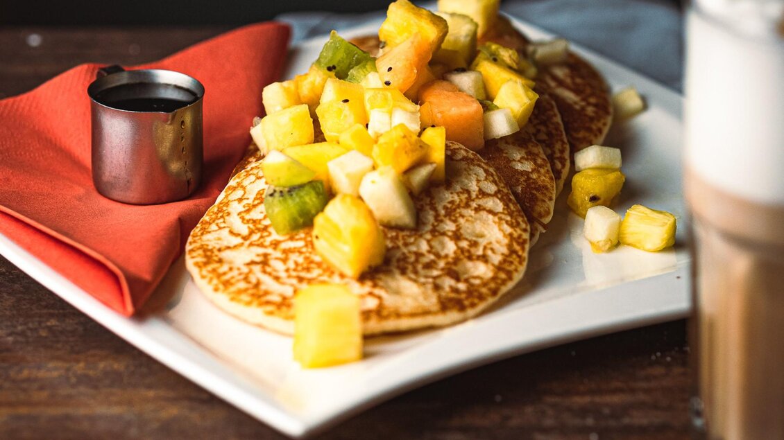 Leckere Pancakes mit frischen Fruchtstücken und einer kleinen Kanne Sirup. Ein rotes Serviette ergänzt die schöne Präsentation. | © Granola