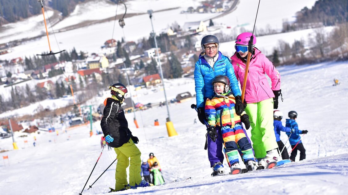 Eine Familie beim Skifahren auf einem sonnigen Hang. Im Hintergrund sind schneebedeckte Berge und ein malerisches Dorf zu sehen. | © Gaaler Skilifte