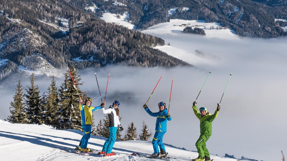 Vier Skifahrer stehen auf einer schneebedeckten Piste und heben ihre Skistöcke in die Luft. Im Hintergrund sind verschneite Berge und eine Wolkendecke zu sehen. | © Gaaler Skilifte