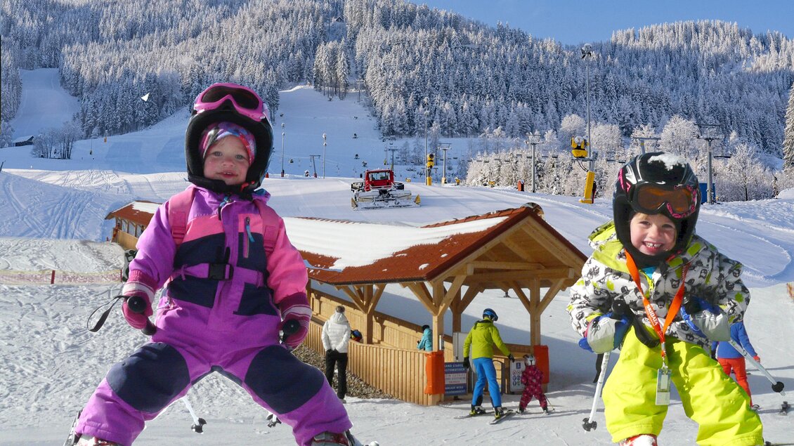 Zwei Kinder auf Skiern stehen fröhlich im Schnee. Im Hintergrund sind verschneite Berge und Skifahrer zu sehen. | © Gaaler Lifte